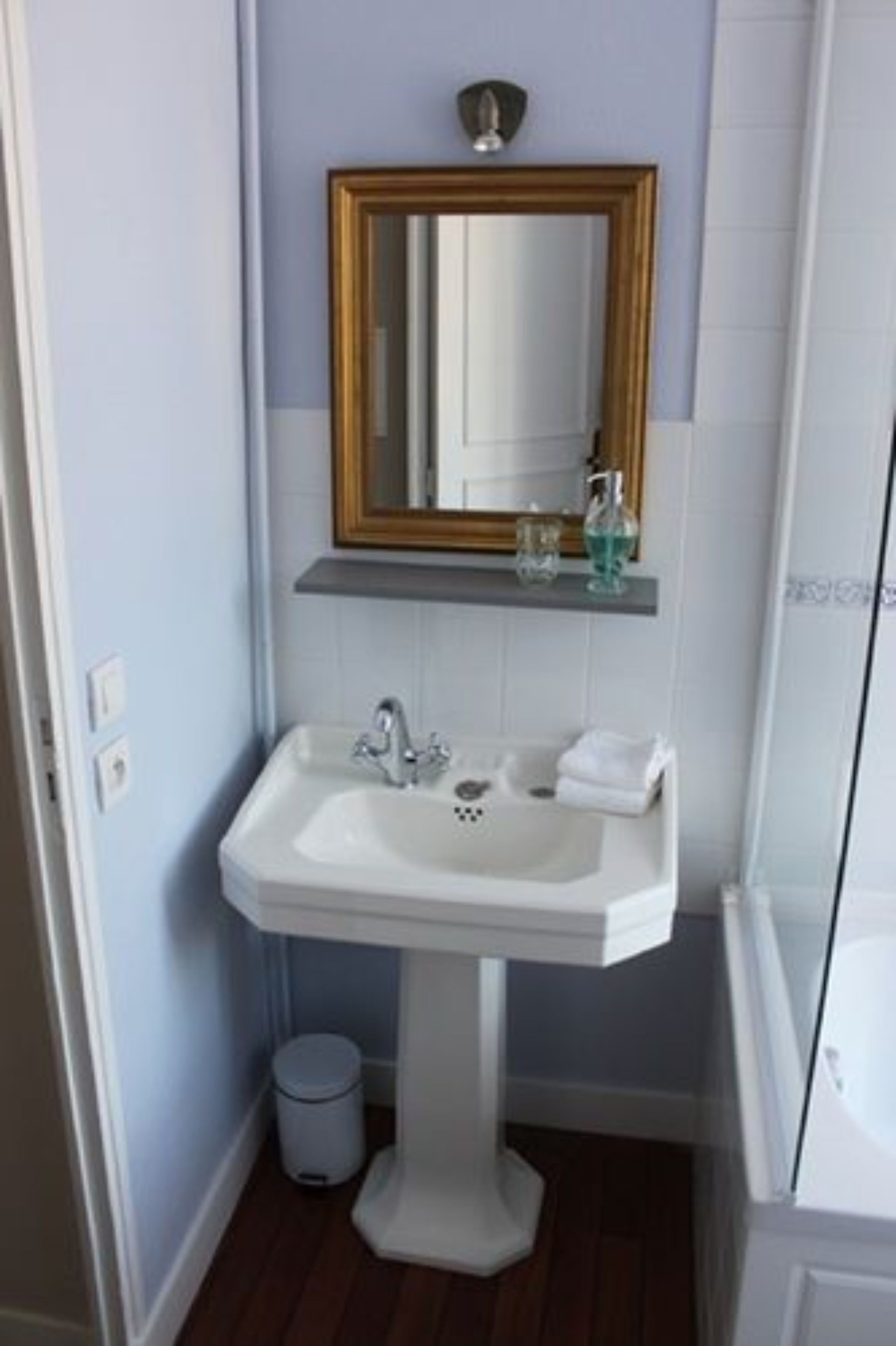 A bathroom with a pedestal sink, a mirror with a gold frame, a small shelf with toiletries, and a trash bin.