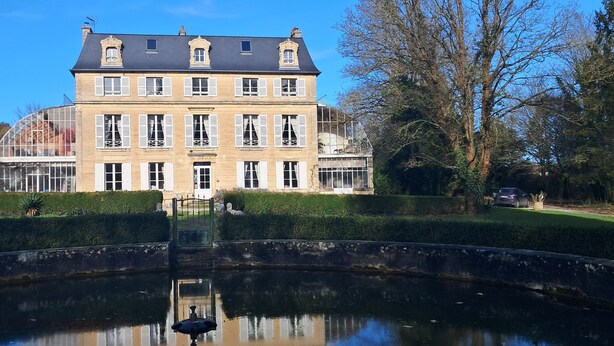 Un grand bâtiment historique doté de nombreuses fenêtres et d’une entrée centrale, entouré d’un jardin bien entretenu et d’un bassin aux eaux calmes et réfléchissantes.