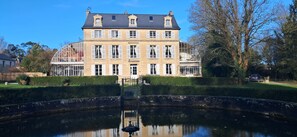 Een groot, historisch gebouw met meerdere ramen en een centrale ingang, omgeven door een verzorgde tuin en een vijver.