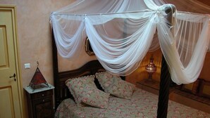 A bedroom with a four-poster bed, floral bedding, a bedside table with a lamp, and a wall-mounted clock. - Le Mazet De La Dame (Barbentane)