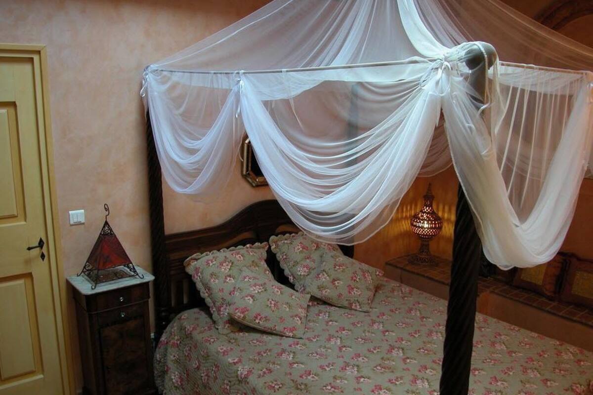 A bedroom with a four-poster bed, floral bedding, a bedside table with a lamp, and a wall-mounted clock.