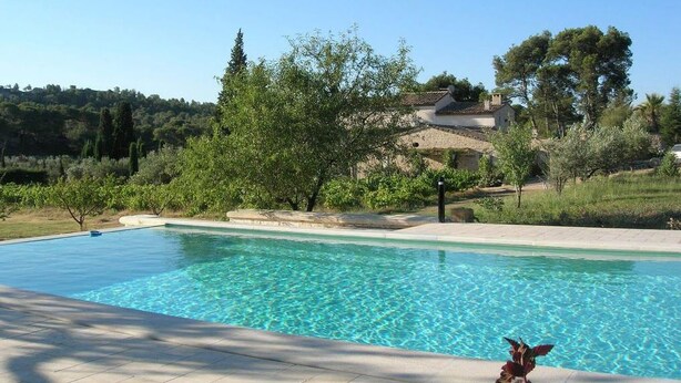 A swimming pool with clear blue water, surrounded by a landscaped garden with trees and shrubs, and a building with a tiled roof in the background. - Le Mazet De La Dame (Barbentane)