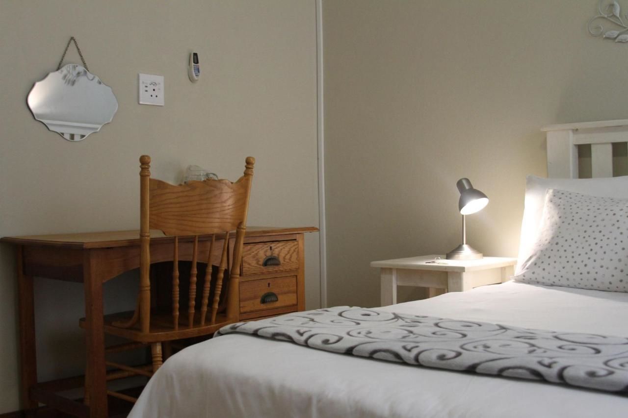 A bedroom with a wooden chair, a bedside table with a lamp, a white bed with a patterned bedspread, and a wall-mounted light fixture.