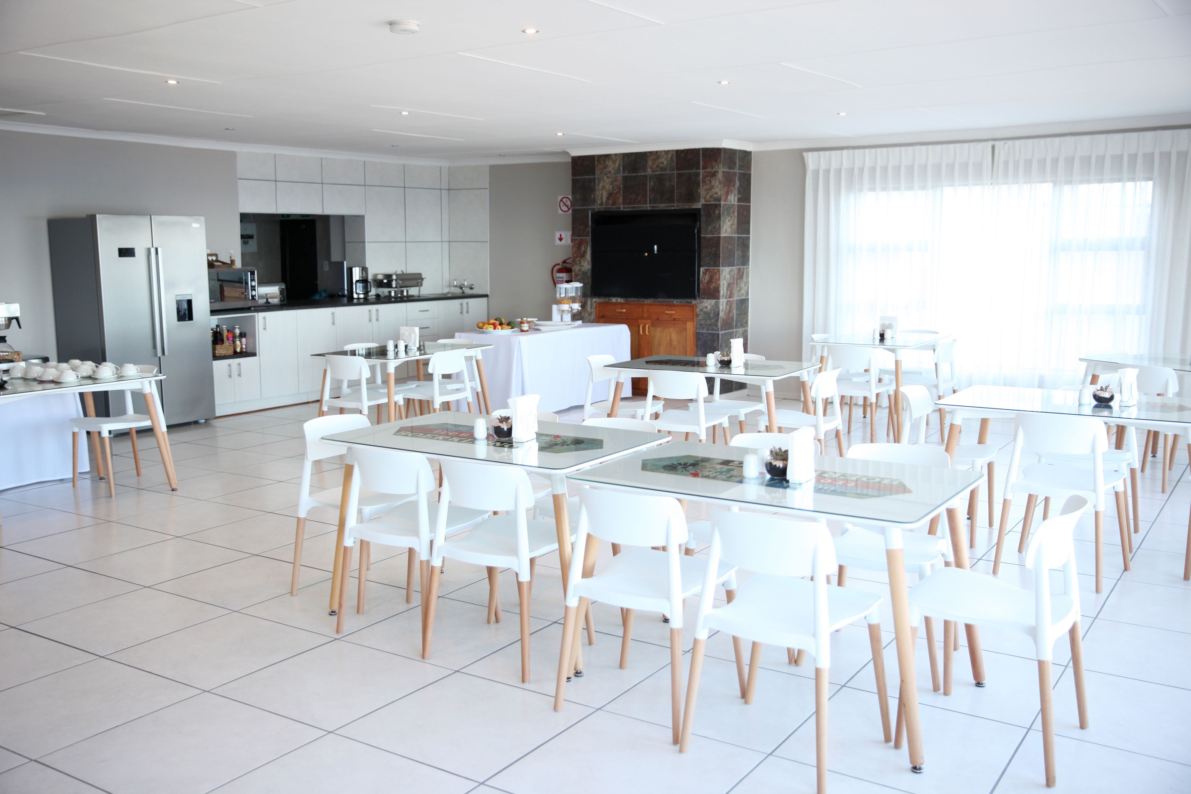 A modern dining area with white chairs and tables, a kitchen in the background, and a large flat-screen TV mounted on the wall.
