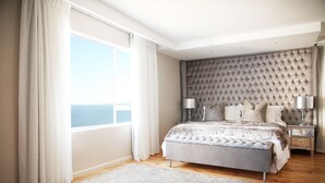 Un dormitorio con una cama grande, un cabecero tapizado, una mesita de noche con lámparas y vistas al mar.