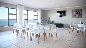 Una sala de estar moderna con una gran mesa de vidrio rodeada de sillas blancas, un televisor de pantalla plana montado en la pared y vistas al océano a través de amplios ventanales.