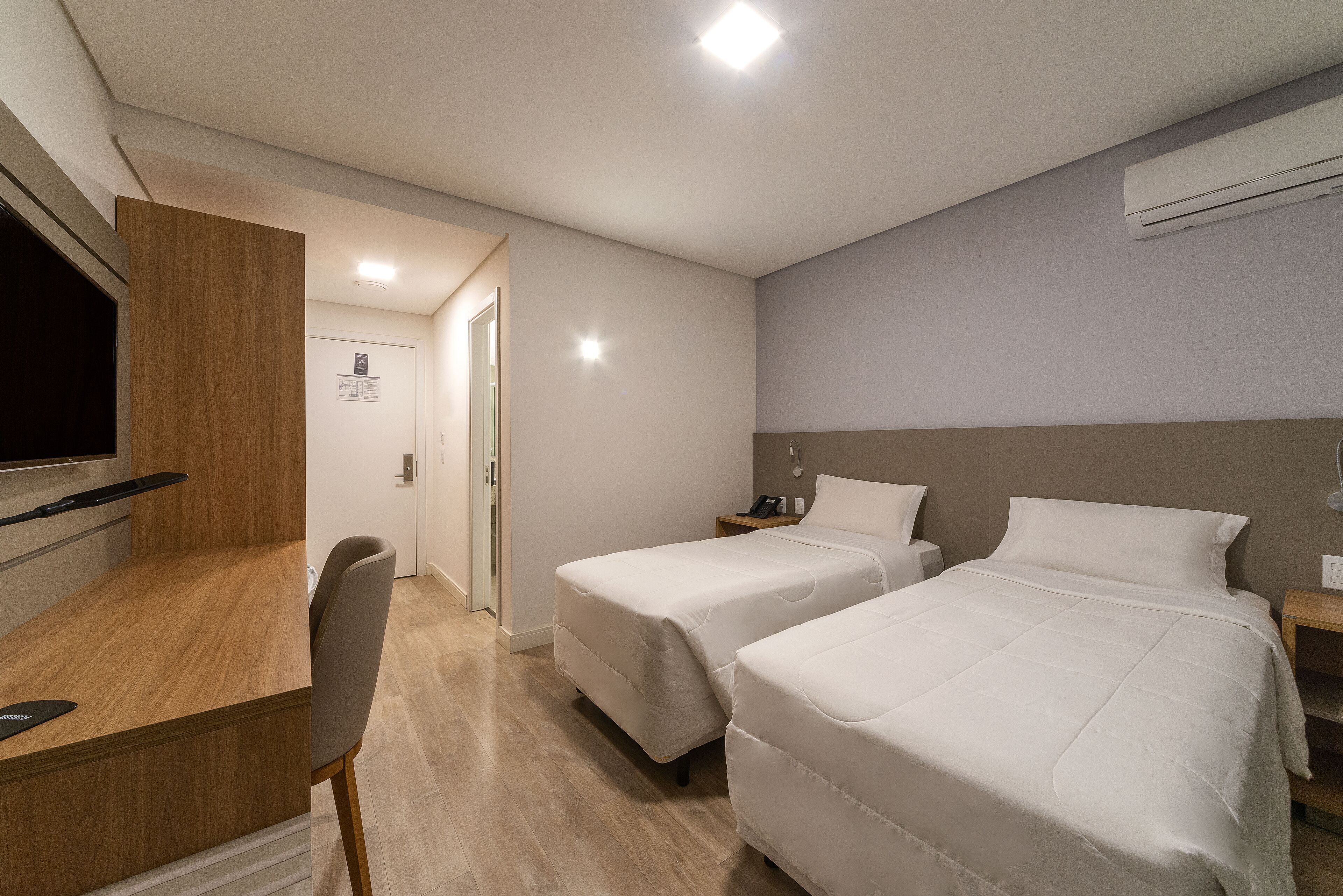 A hotel room with two beds, a desk, a chair, a TV, and an air conditioning unit.