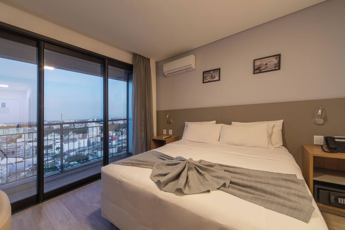 A hotel room with a large bed, a view of the city through floor-to-ceiling windows, a bedside table with a lamp, and an air conditioning unit on the wall.
