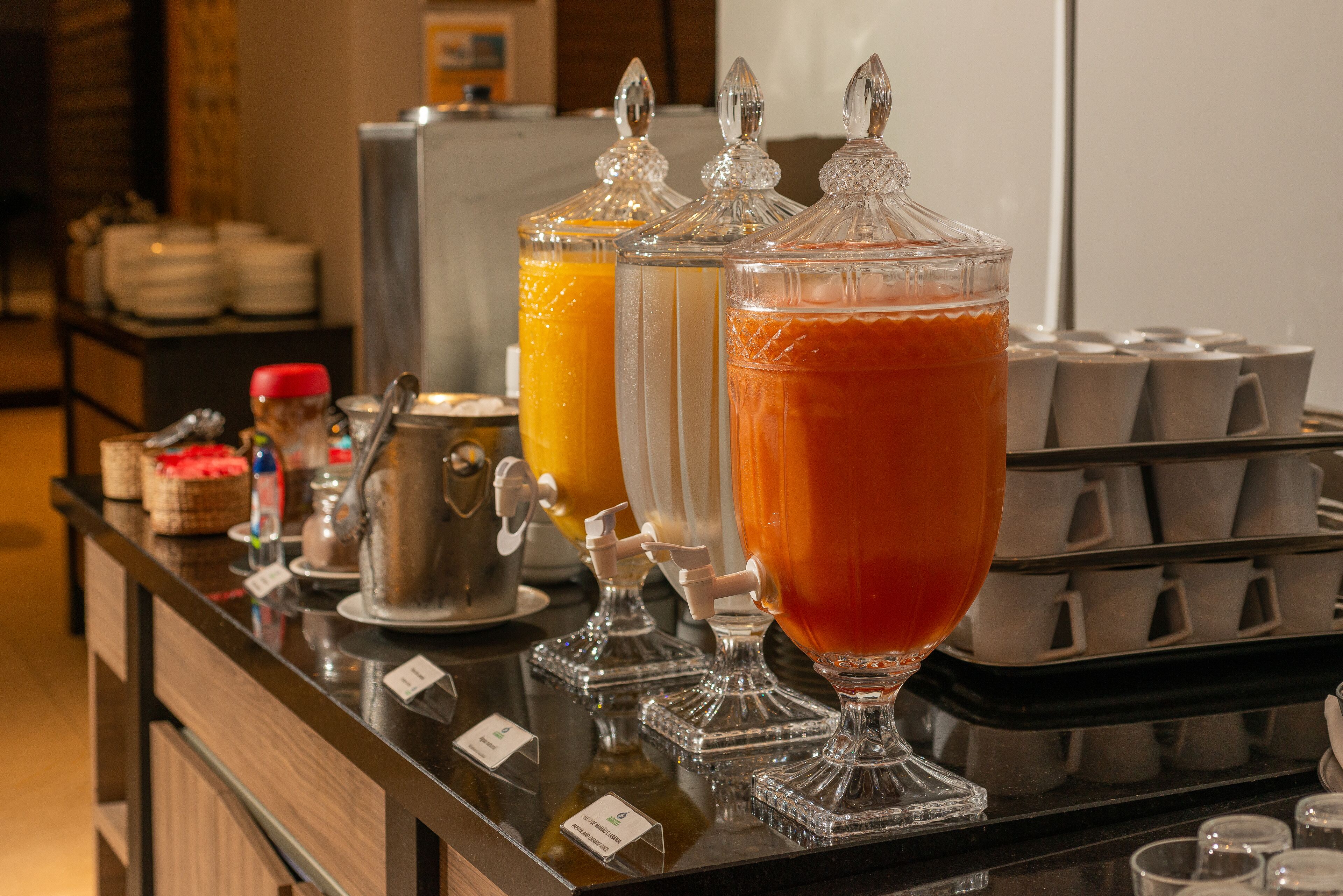 Una barra de desayuno con dispensadores de vidrio transparente para jugo de naranja y té, una máquina de café y una bandeja de vasos desechables.