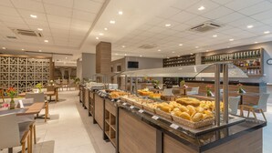 A modern cafe with a long counter displaying various baked goods and pastries, wooden tables and chairs, and a wall of neatly arranged bottles. -  Laghetto Rio Grande (Rio Grande)