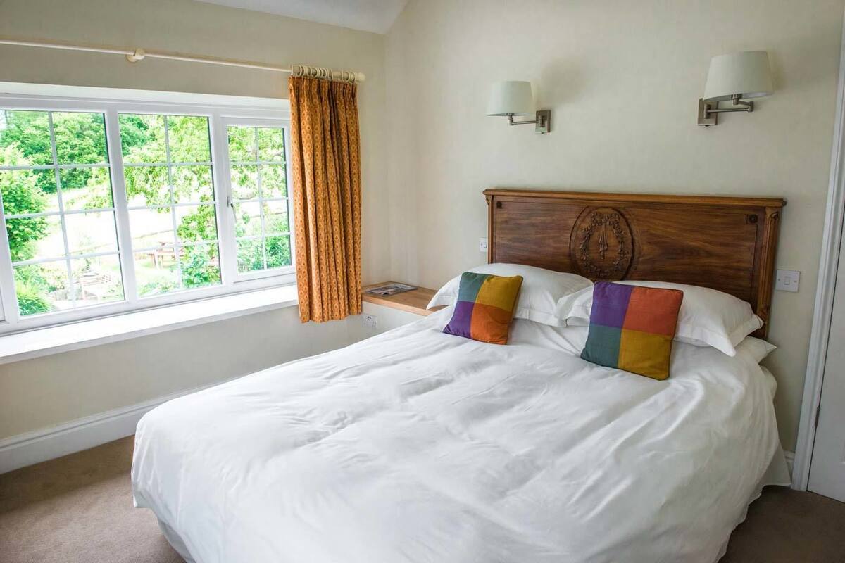 A neatly made bed with white linens and colorful pillows, a wooden headboard, two wall-mounted lamps, and a window with curtains.