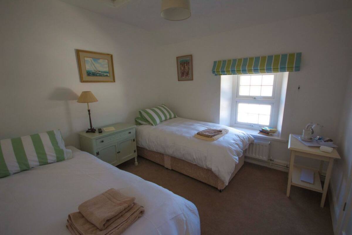 A bedroom with two beds, a nightstand, a lamp, a window with a striped valance, and a small table with a pitcher.