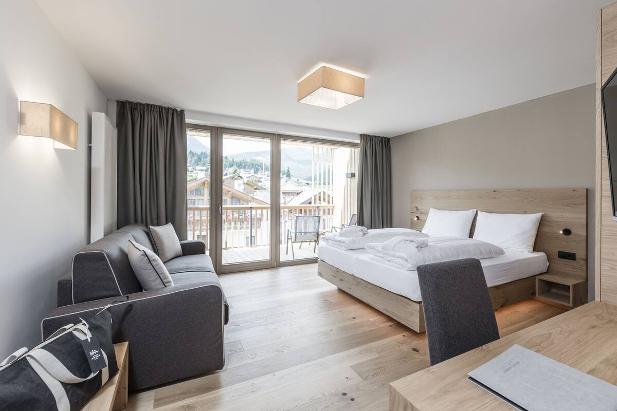 A modern hotel room with a bed, a sofa, a desk, and a view of the city through large windows.