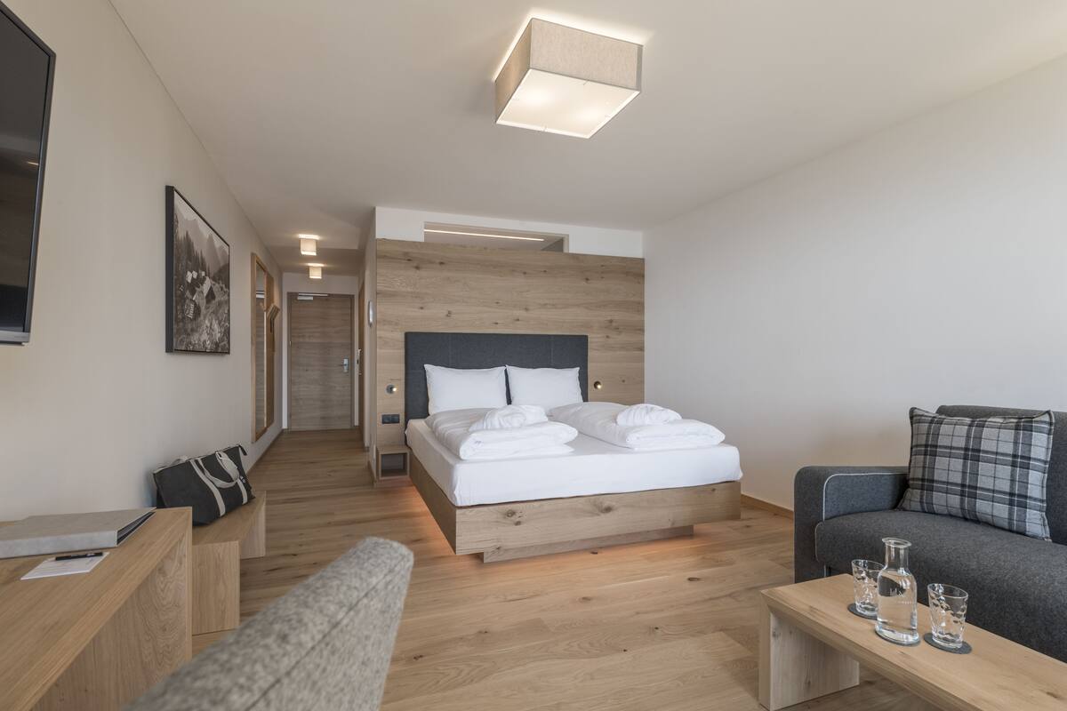 A modern hotel room with a wooden bed, a sofa, a desk, and a small table with glasses.