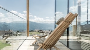 A wooden lounge chair on a balcony with a glass railing, overlooking a mountainous landscape and a village below. - Hotel Ambet (Rio di Pusteria)