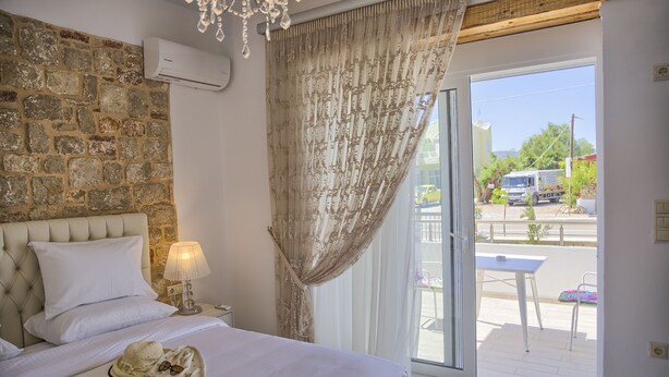 A bedroom with a stone wall, a bed with white linens, a bedside lamp, and a view of the outside. - G&K Stone Suites (Rhodes)