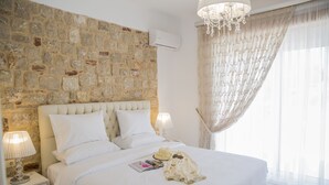 A bedroom with a stone wall, a large bed with white bedding, a chandelier, and a window with sheer curtains. - G&K Stone Suites (Rhodes)