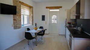 A compact kitchen with a dining area, stone wall, wooden beams, and a TV mounted on the wall. - G&K Stone Suites (Rhodes)