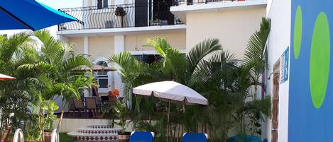 Un área de piscina con sillones de descanso y sombrillas, un edificio de varios pisos con balcones, y un cielo azul despejado.