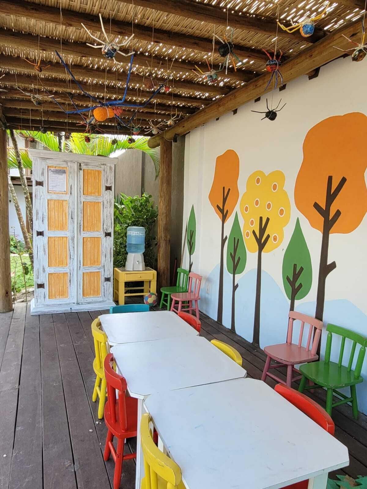An outdoor patio area with a long white table, colorful chairs, and a mural of trees on the wall.