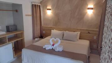 Habitación de hotel con cabecera de madera, dos lámparas de noche, cama con colcha blanca y almohadas, mesita de noche con teléfono y pequeño escritorio con microondas.