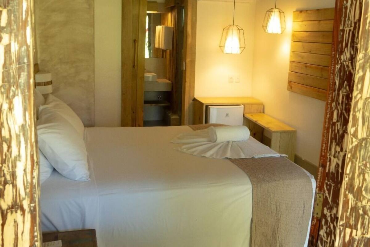 A bedroom with a bed, bedside tables, a nightstand, a wooden headboard, a hanging light fixture, and a mirror with a wooden frame.