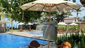 Un espace aménagé au bord de la piscine, avec des chaises et des tables en bois sous des parasols, une fontaine en pierre et une vue sur l’océan.