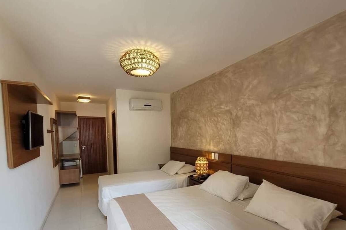 A hotel room with two beds, a television mounted on the wall, a ceiling light fixture, and an air conditioning unit.