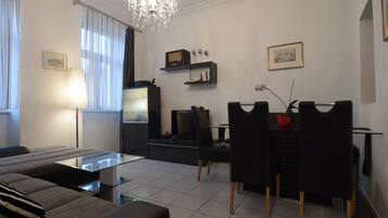 A modern living room with a sofa, a glass coffee table, a dining area with chairs, a chandelier, and a TV.