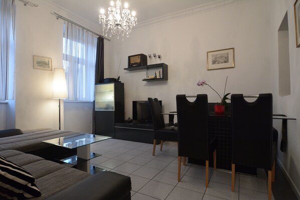 A modern living room with a sofa, a glass coffee table, a dining area with chairs, a chandelier, and a TV.