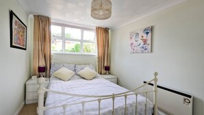 Un dormitorio con cama blanca, cortinas color beige, una ventana con vistas, una mesita de noche con lámpara y un cuadro en la pared.