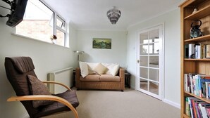 Una habitación acogedora con un sillón de cuero marrón, un sofá beige con almohadones, una estantería repleta de libros, una araña y una puerta que da a otra habitación.