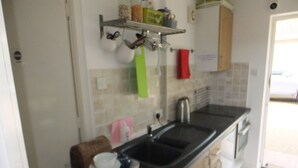 Una cocina pequeña con un lavamanos, una cafetera y un tostador sobre la encimera. Hay una estantería encima del lavamanos con varios objetos.