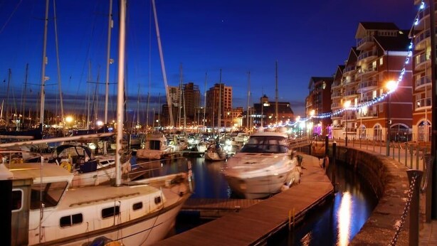 Un puerto deportivo al atardecer con barcos amarrados, un canal de agua y edificios con luces.