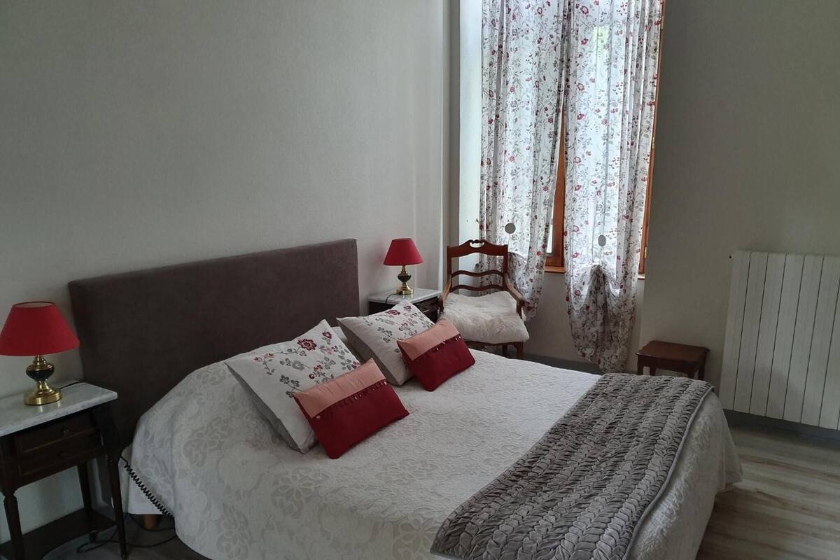 Une chambre à coucher bien aménagée, avec un grand lit, deux tables de chevet, une chaise et une fenêtre ornée de rideaux à motifs floraux.