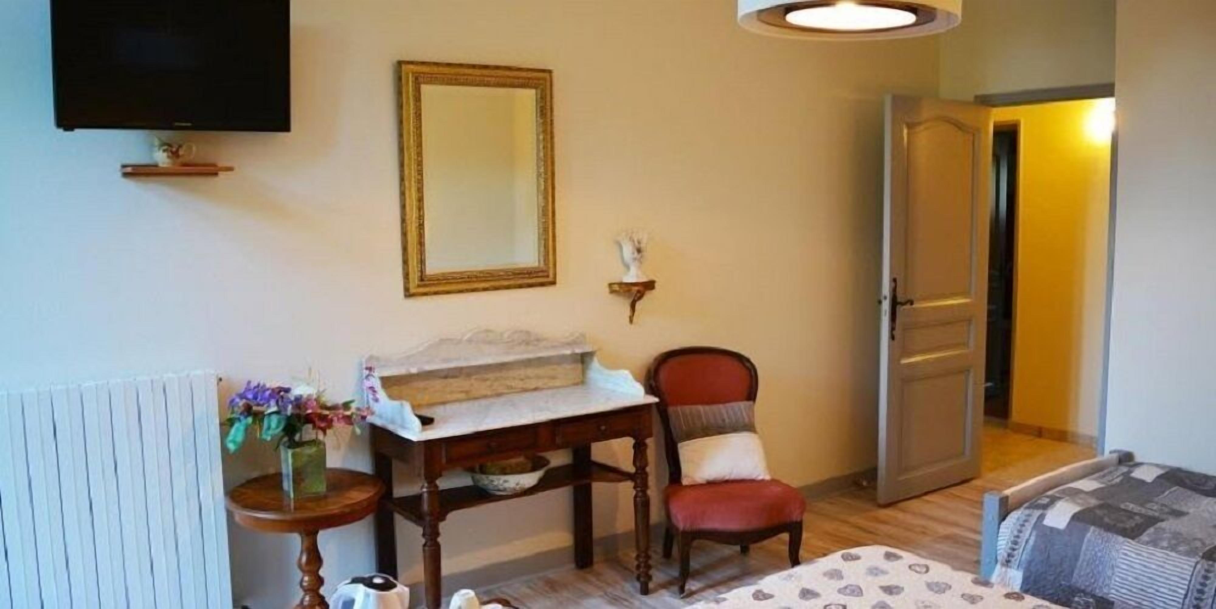 A room with a wooden chair, a small table with a vase of flowers, a mirror on the wall, and a bed with patterned bedding.