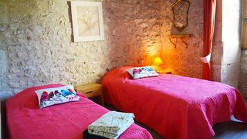 A room with two beds, each with a red bedspread and a pillow with a bird design. There is a small wooden nightstand with a lamp between the beds. The walls are textured and one wall has a framed picture.
