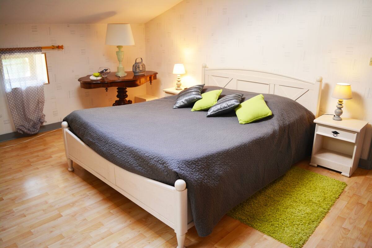 Un lit bien fait, avec une couverture grise et des coussins verts, une table de chevet en bois avec une lampe, et un tapis vert sur un plancher en bois.