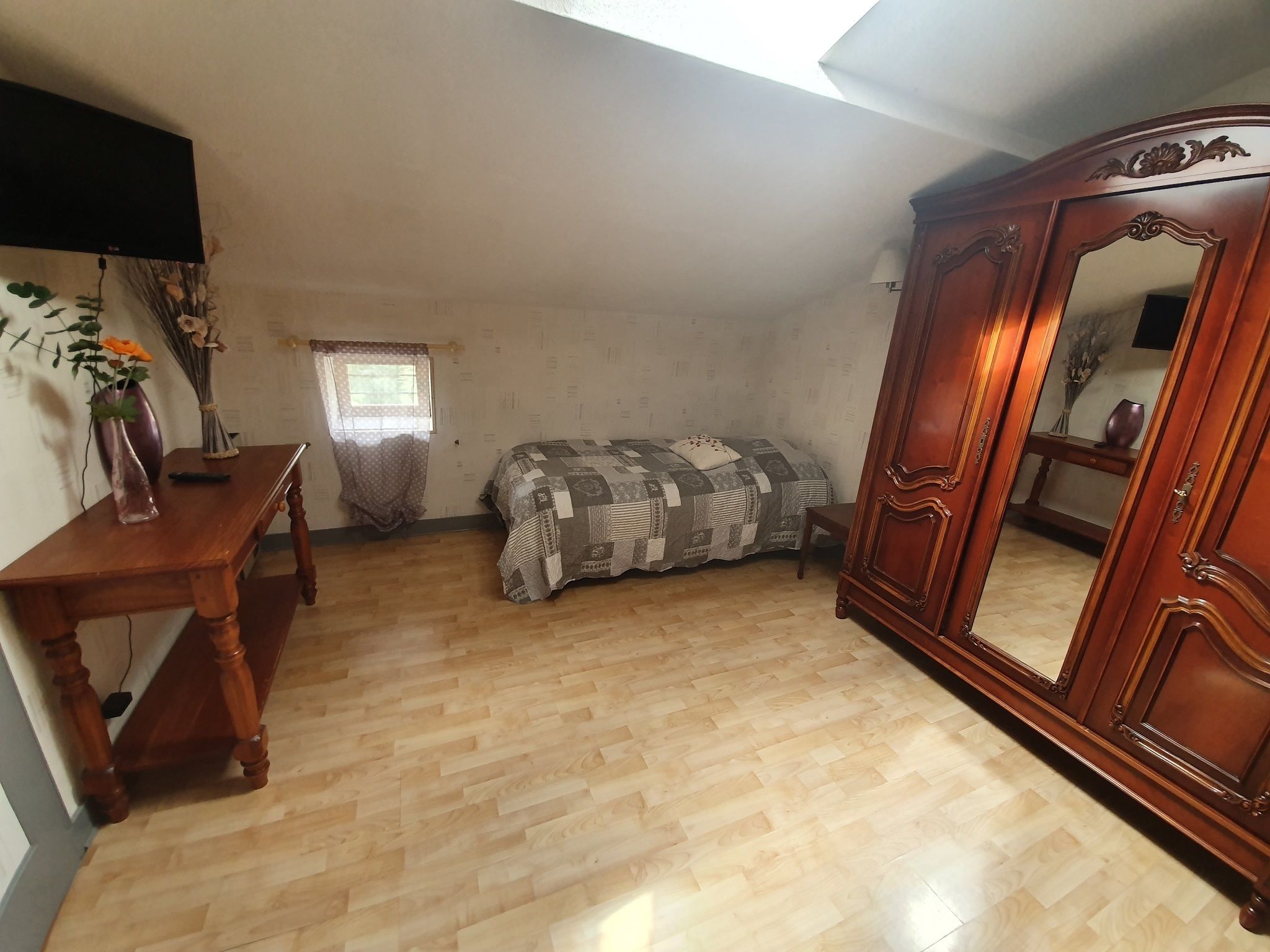 Un dormitorio con suelo de madera, una cama con manta estampada, un armario de madera, un escritorio con un florero y una ventana con cortinas.