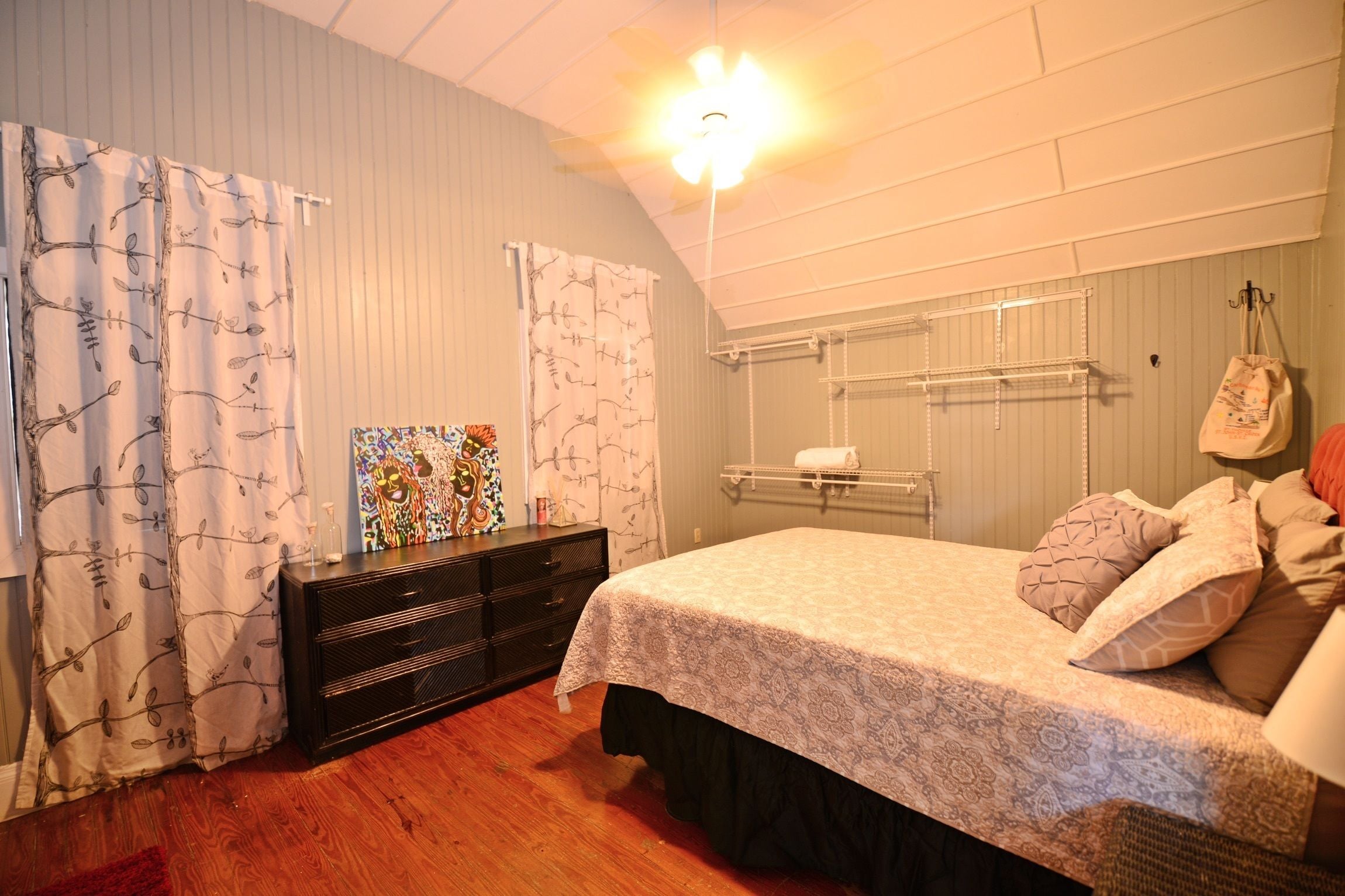 A bedroom with a bed, a dresser, a shelf with towels, and a hanging bag.