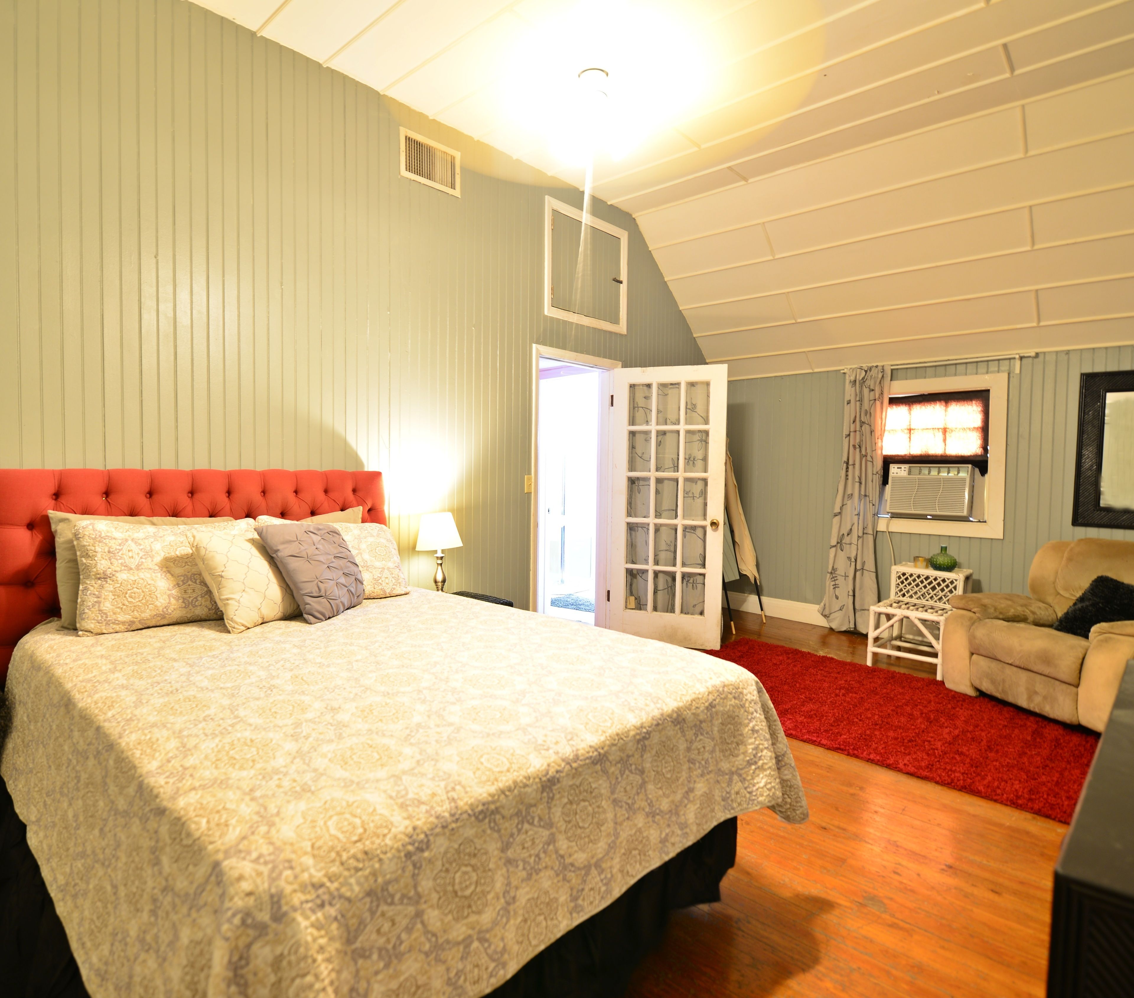 A bedroom with a large bed, a red upholstered headboard, a red carpet, a small white side table, and a beige armchair.