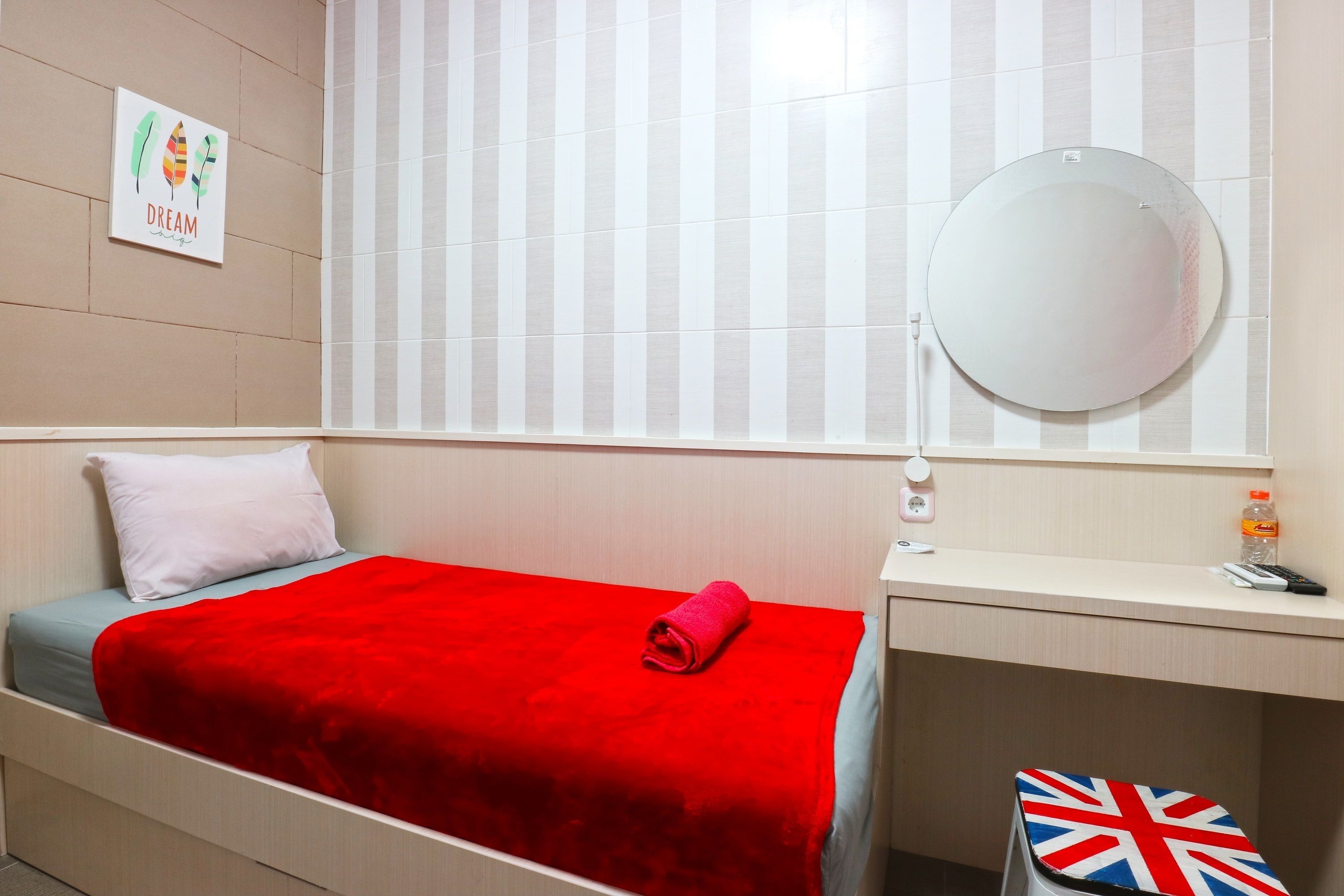 Quarto de hotel com uma cama, um cobertor vermelho, uma mesa pequena com uma garrafa, um espelho e uma parede com a placa 'DREAM'.