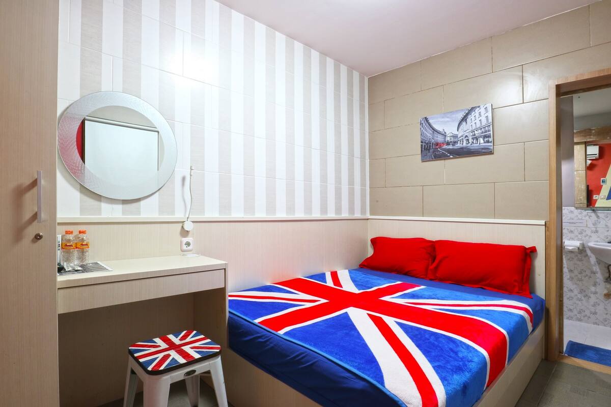 A hotel room with a bed featuring a Union Jack blanket, a round mirror on the wall, a small stool with a Union Jack design, and a picture of a city street on the wall.