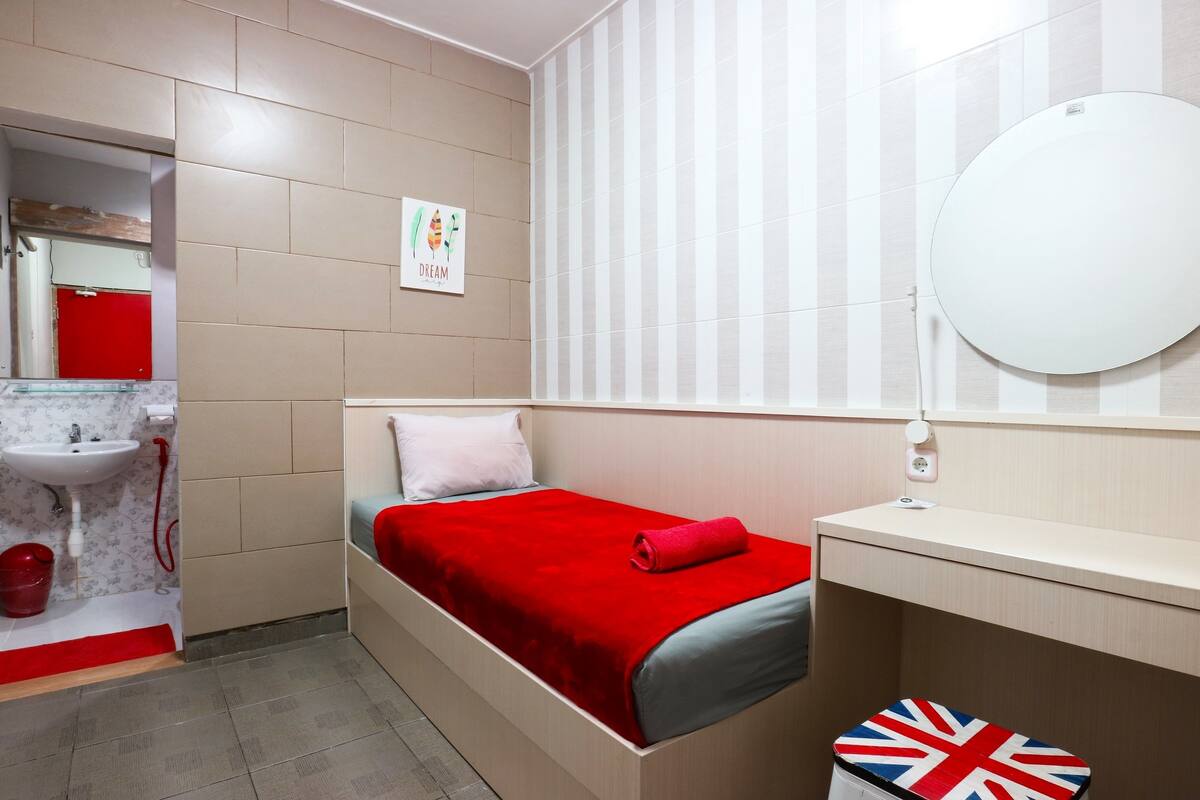 A compact room with a bed, a small bedside table, a mirror, a red towel, a red bucket, and a sink with a red cabinet.