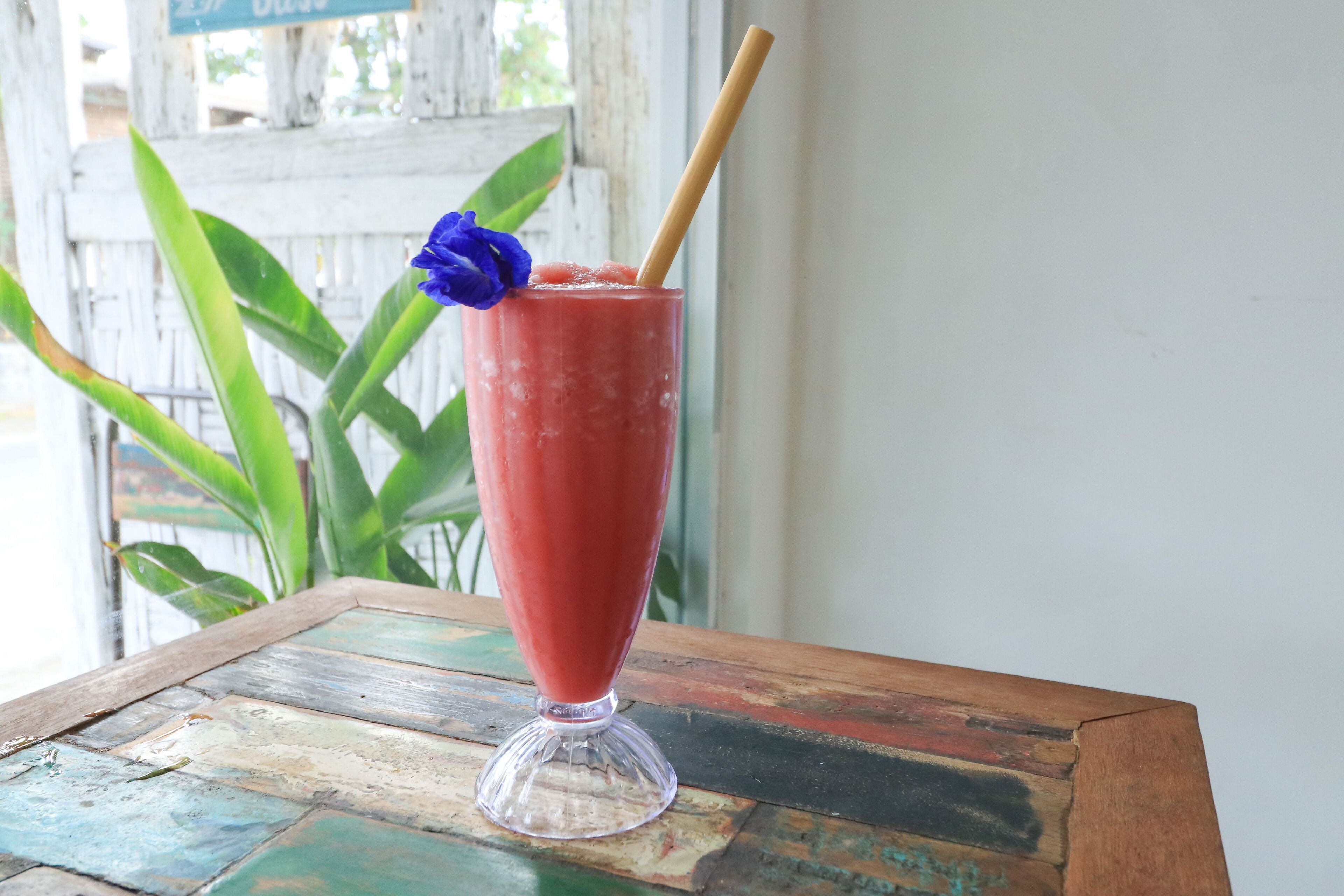 A glass of red beverage with a blue flower and a straw on a rustic wooden table.