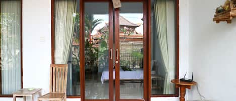 Quarto com porta de correr de vidro que dá para uma área externa coberta, uma cadeira de madeira, uma mesa pequena e uma estante decorativa.