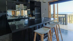 A modern kitchen with a large island, bar stools, and a view of the ocean through a balcony.