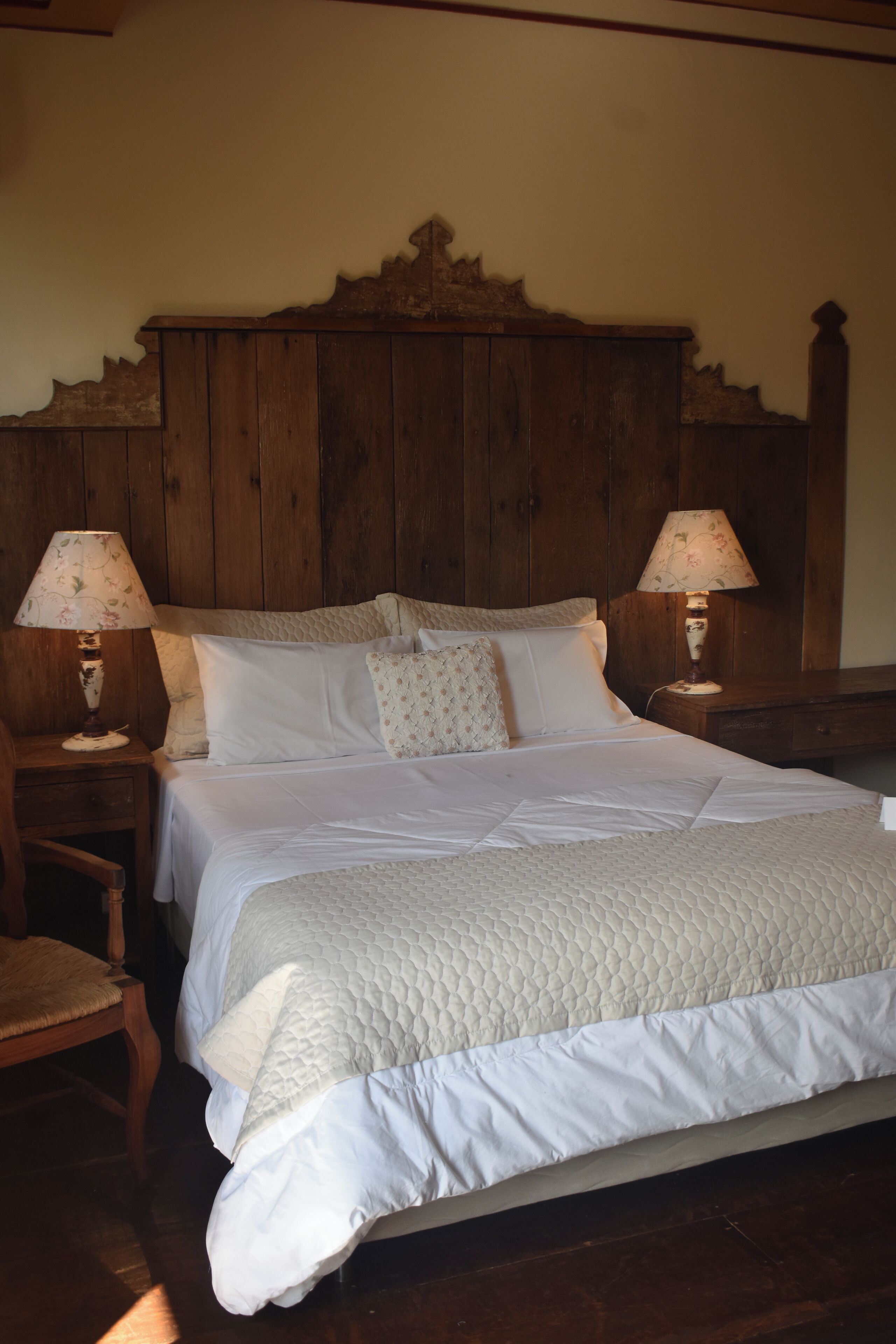 Een slaapkamer met een houten hoofdbord, een bed met wit beddengoed, twee nachtkastjes met lampen en een houten stoel.