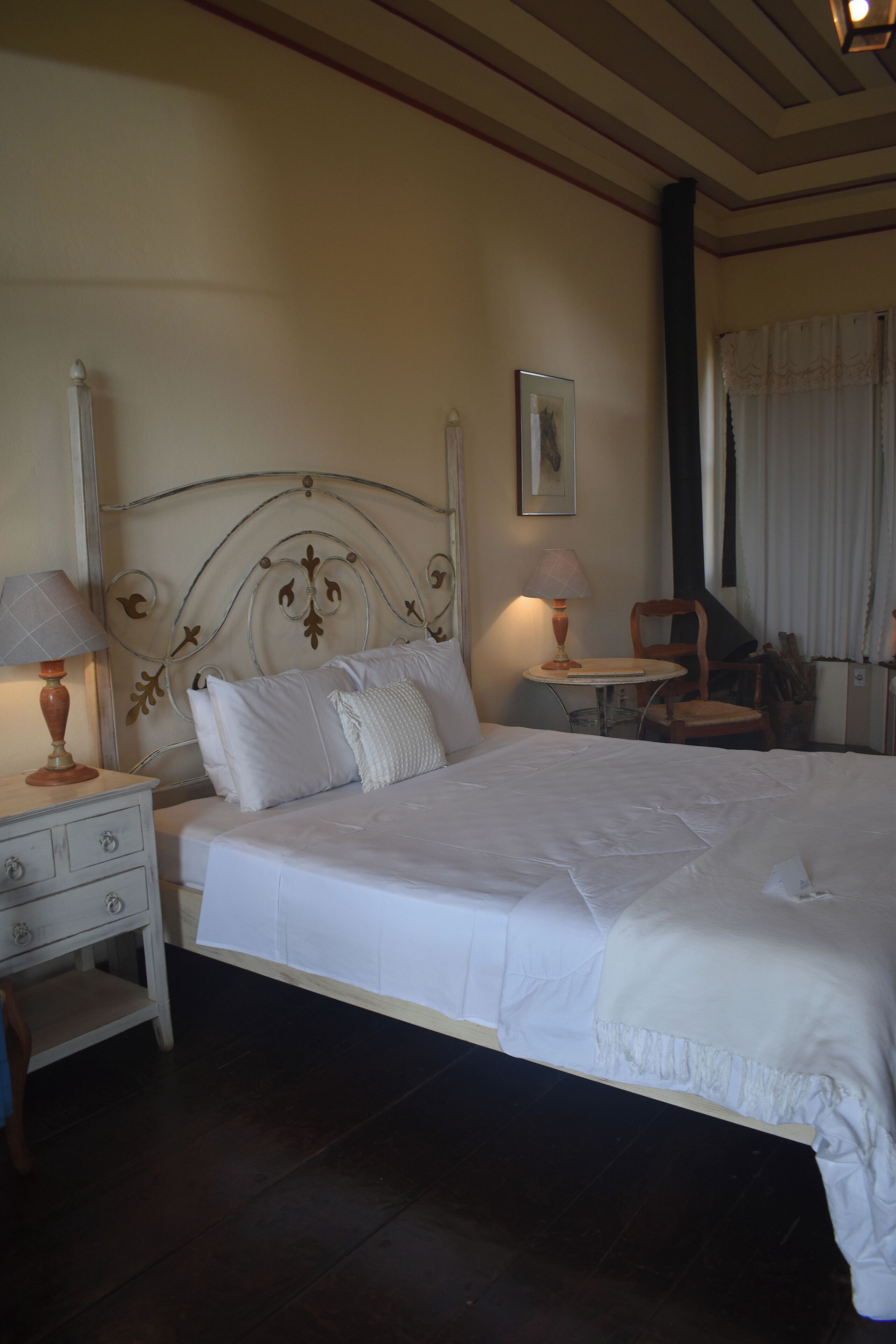 A neatly made bed with white linens, a bedside table with a lamp, and a chair in a room with wooden flooring and a decorative ceiling.