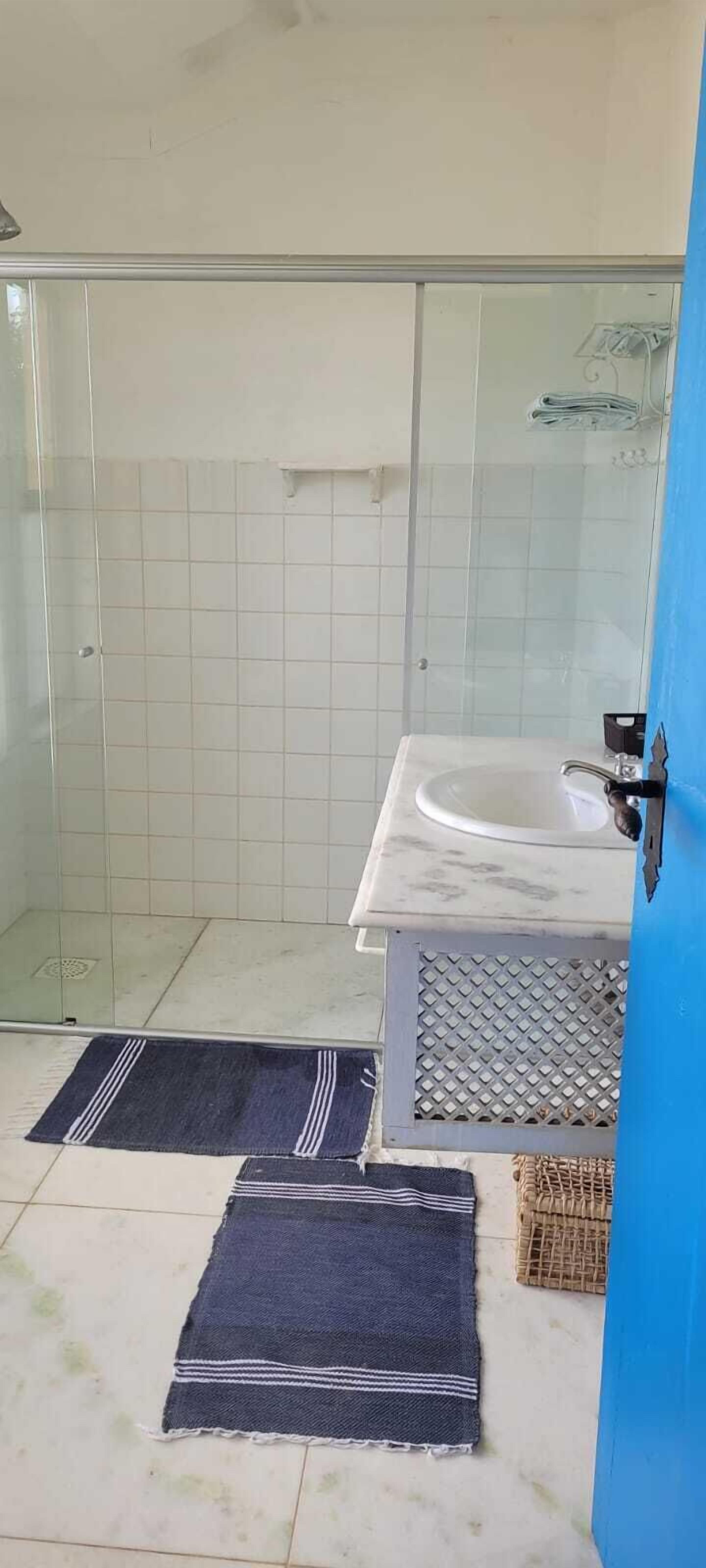 Casa de banho com cabine de chuveiro em vidro, lavatório branco e dois tapetes escuros no chão.